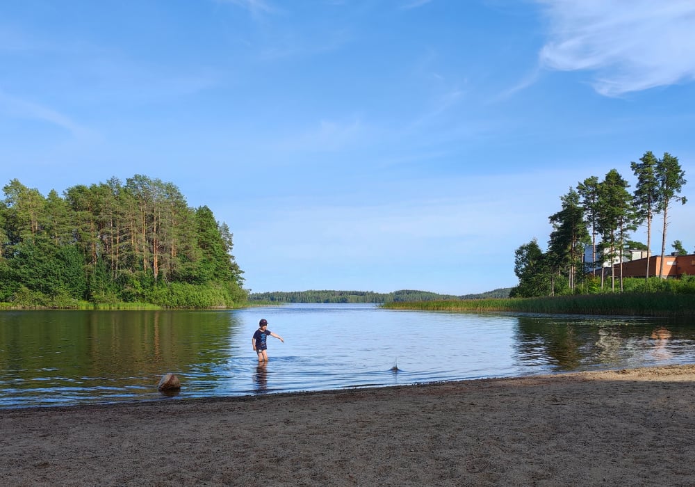 Kuvassa uimaranta kesäisenä päivänä ja poika kahlaamassa vedessä